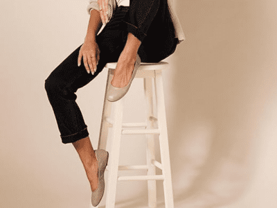 A woman is sitting on a stool wearing black pants and nude ballet flat sandals.