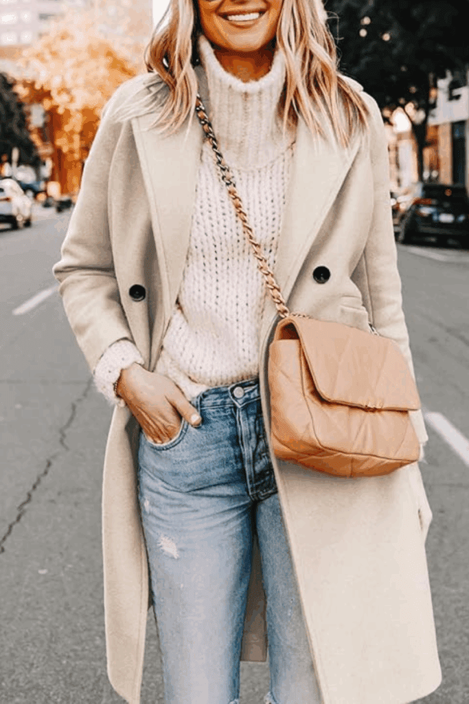 A smiling woman wearing jeans, a cream chunky turtleneck and a Beige wool coat. She has a brown crossbody bag with gold detail.